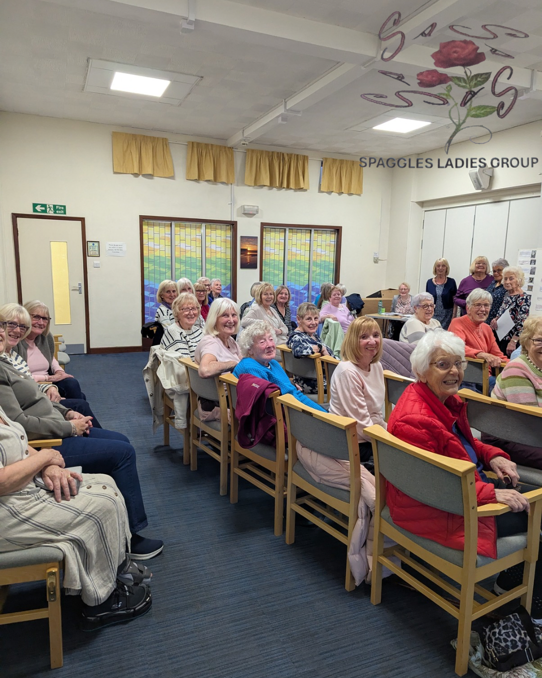 A large group of older women are seated in rows of chairs in a community hall, smiling and looking towards the camera. The bright room features colourful stained-glass-style window panels and cream curtains. Several women stand at the back of the room, while others chat and enjoy the gathering. The image includes the Spaggles Ladies Group logo in the top-right corner.