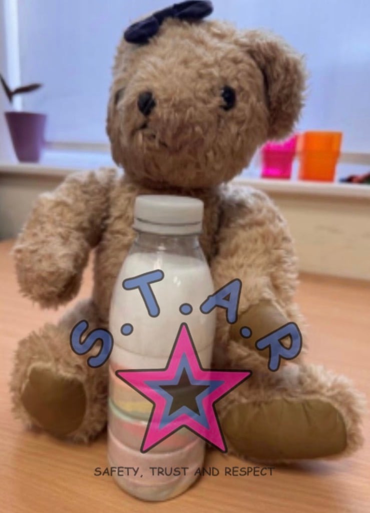 A well-loved brown teddy bear sits on a wooden table holding a baby bottle filled with milk. The teddy wears a dark bow on its head and is positioned in front of a softly blurred indoor background. Overlaid on the image is the S.T.A.R. logo, featuring a pink and blue star and the words “Safety, Trust and Respect.”