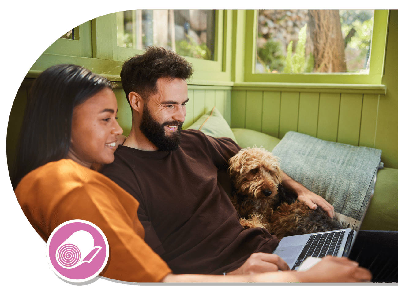 A man and a woman sitting side by side on a couch, looking at a laptop, with a small brown dog nestled between them in a warmly lit indoor setting.