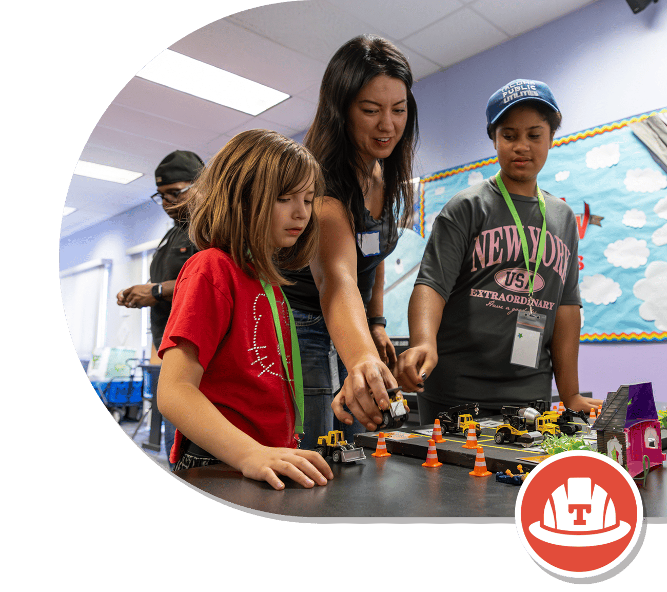 A woman and two children work together at a table with toy construction vehicles and colorful miniature houses, in a bright classroom setting.