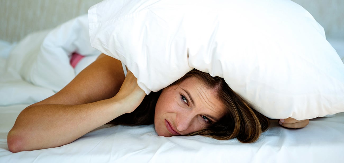 Woman laying on a bed with a pillow over her head looking frustrated