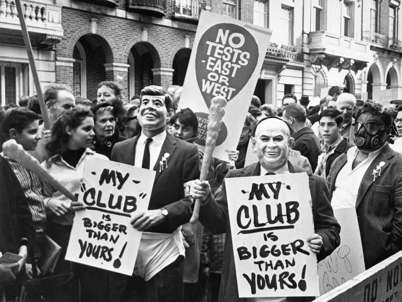A crowded street protest with people holding signs reading MY CLUB IS BIGGER THANYOURS and wearing masks including caricatures and a gas mask one sign says NO TESTS EAST OR WEST