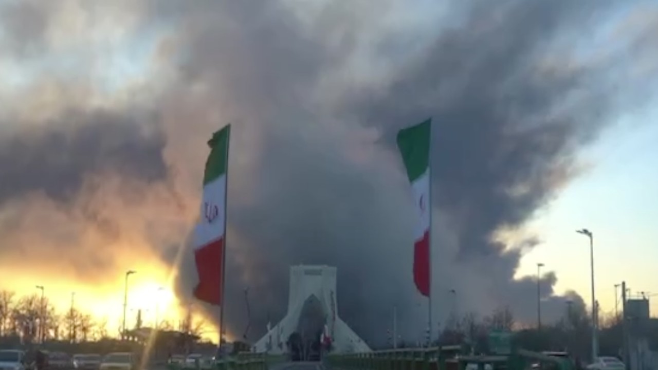 A huge cloud of smoke rises behind a monument framed between two Iranian flags