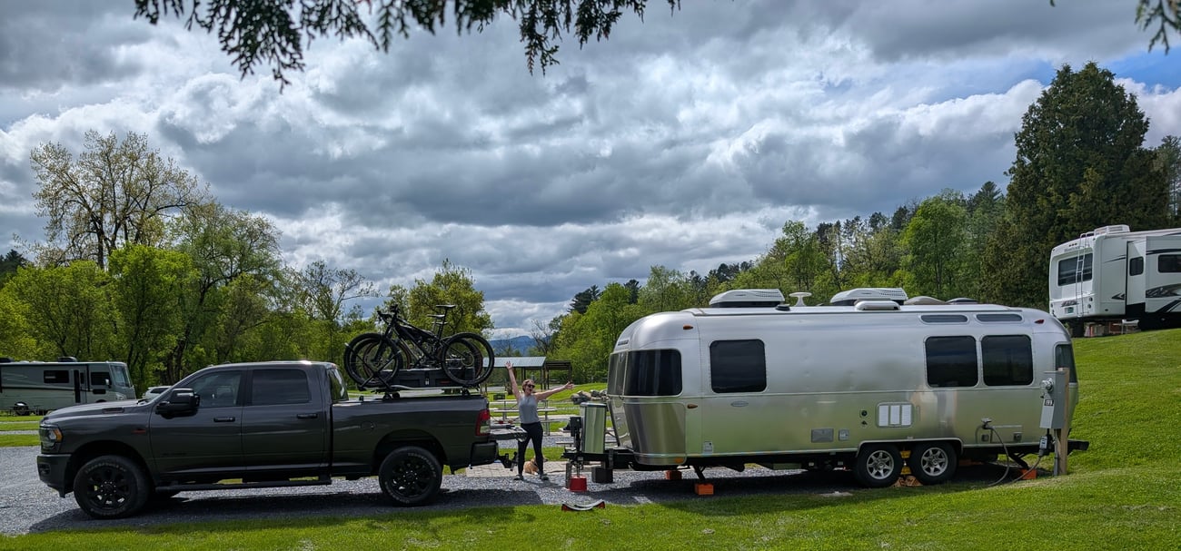 Truck towing Airstream travel trailer at a green campsite under cloudy skies.