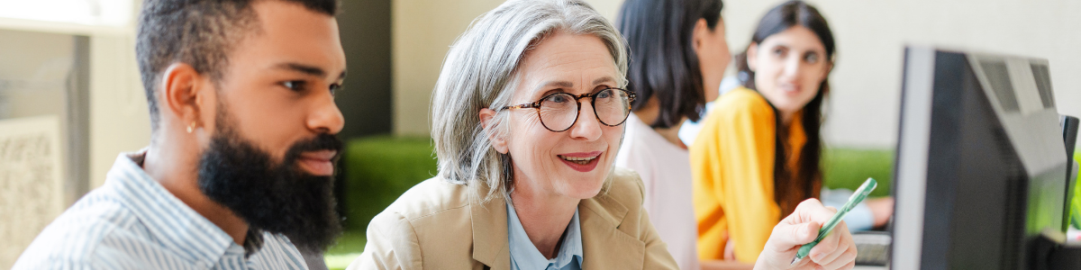 Two people sit in front of a computer, with one gesturing towards the screen, and other people sitting in the background. 