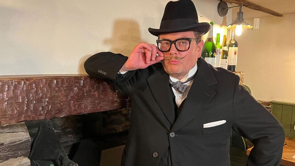 A man in a suit and hat leans casually against a fireplace, exuding a sophisticated and relaxed demeanor. A man in a suit and hat leans casually against a fireplace, exuding a sophisticated and relaxed demeanor.
