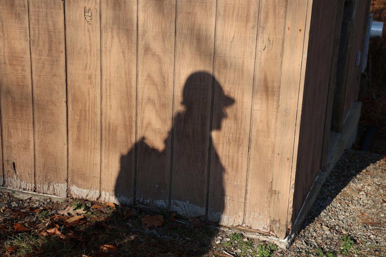 A high-contrast shadow of a wildland firefighter wearing a fire-rated hard hat and a fire pack is cast against the side of a weathered, light-brown wooden building. The shadow is sharp and clear, silhouetted on the vertical wood siding, with dry autumn leaves and gravel visible on the ground below.