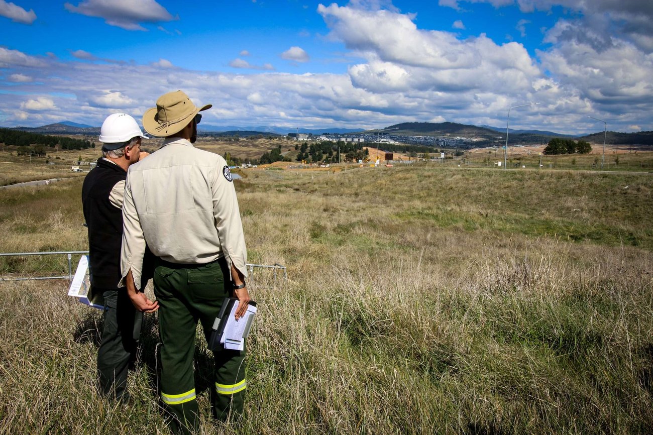 Two land management professionals stand in a grassy field, viewed from behind as they look out toward a distant valley and mountain range. One individual wears a white hard hat and black vest, while the other wears a tan wide-brimmed hat and a uniform with green tactical pants, holding a clipboard. The landscape features rolling dry hills and a small town in the distance under a bright blue sky filled with scattered white clouds.