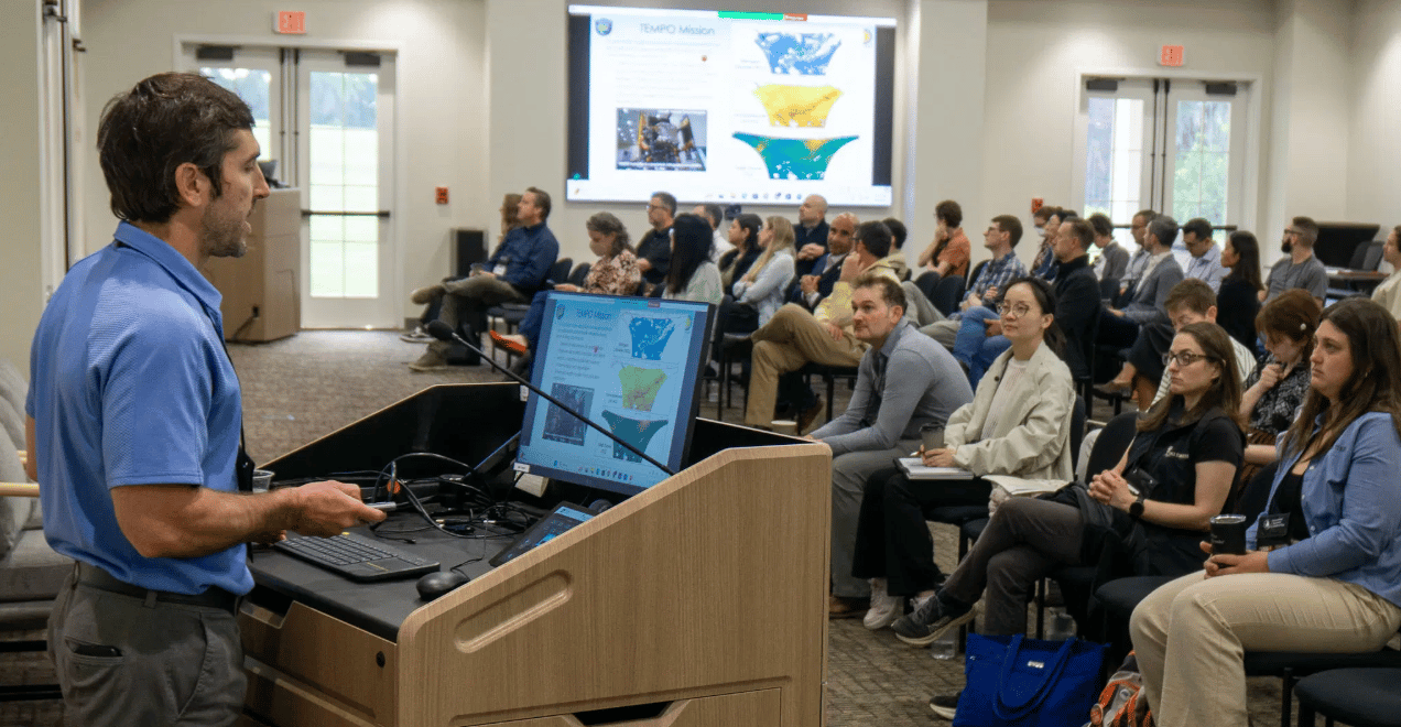 A man in a blue polo shirt stands at a wooden podium, delivering a presentation to a seated audience in a conference room. He is looking toward a large screen displaying colorful heat maps and technical data. The audience, composed of diverse professionals, is listening attentively, with some taking notes and others looking at the presenter. A smaller monitor on the podium mirrors the slides being shown to the room.