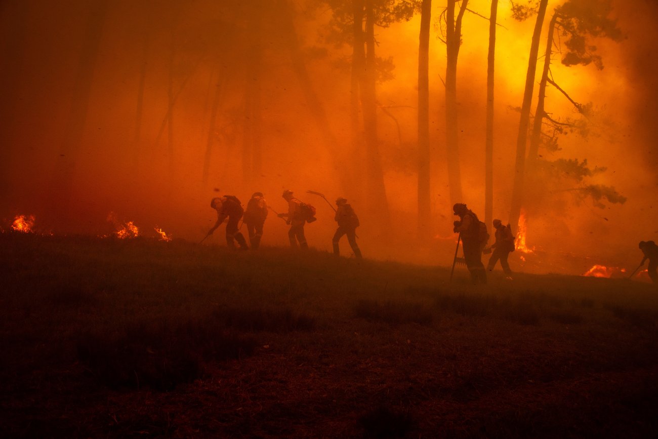 A group of silhouetted wildland firefighters using hand tools to manage a fire line in a dense, smoky forest. The scene is illuminated by an intense, glowing orange light from active flames in the background, highlighting the hazy and dangerous conditions of the fire environment.