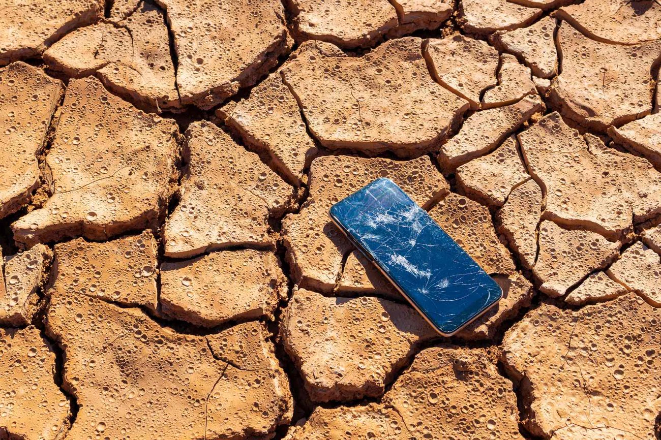 Photo shows a smartphone with a broken screen lying on cracked dry ground