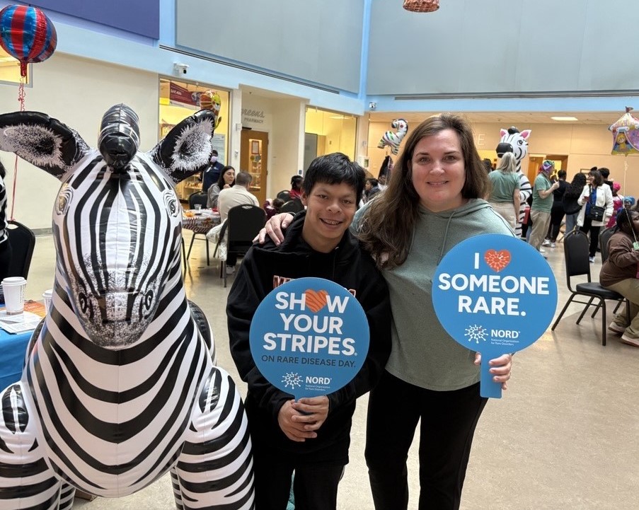 Photo of a parent and child holding signs that say "Show Your Stripes" and "I Love Someone Rare" next to a life-size inflatable zebra