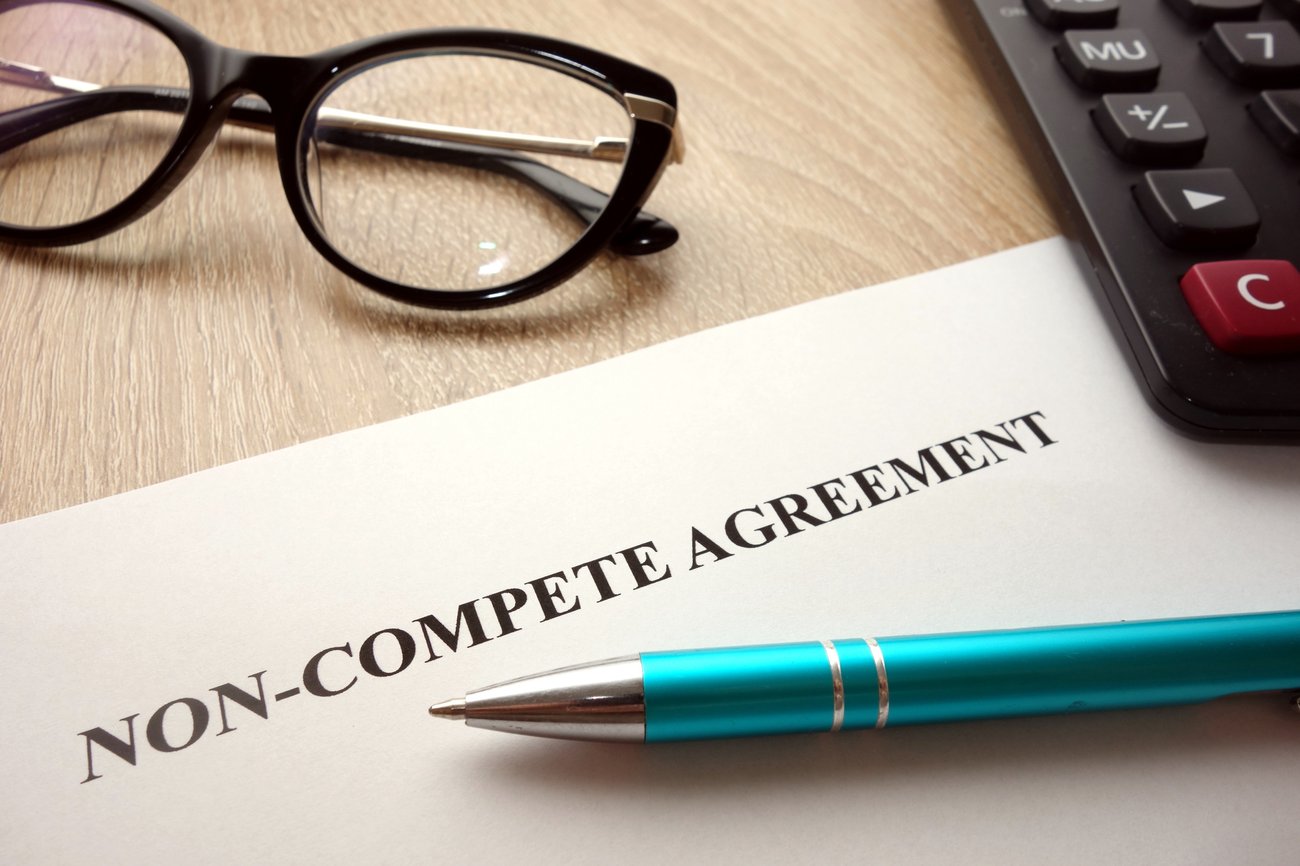a piece of paper reading: "Non-Compete Agreement," surrounding it, a pair of glasses and a pen
