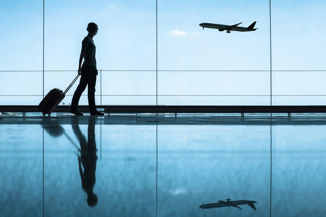 Person with suitcase looking out airport window to see airplane taking off, blue hues.
