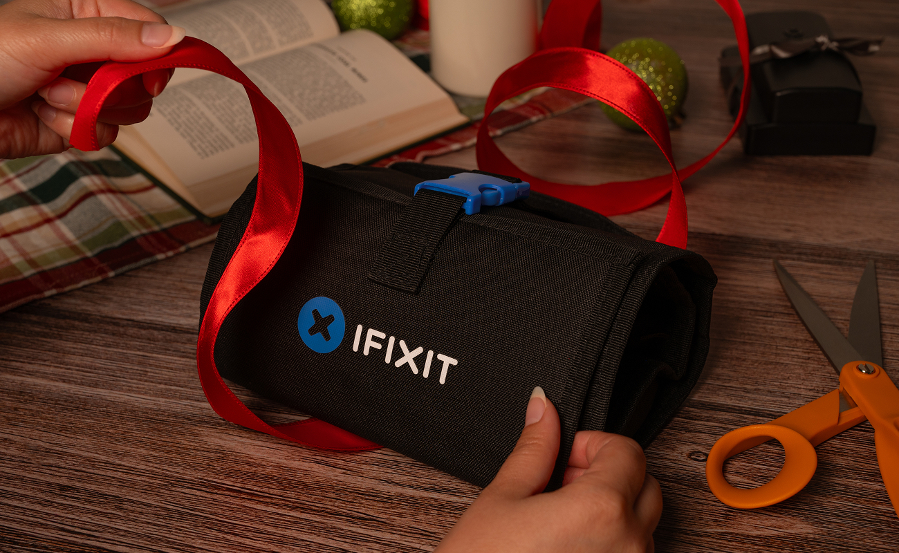 Close-up of hands tying a red ribbon around an iFixit FixHub Toolkit on a wooden table, with holiday décor, an open book, and gift-wrapping tools in the background.