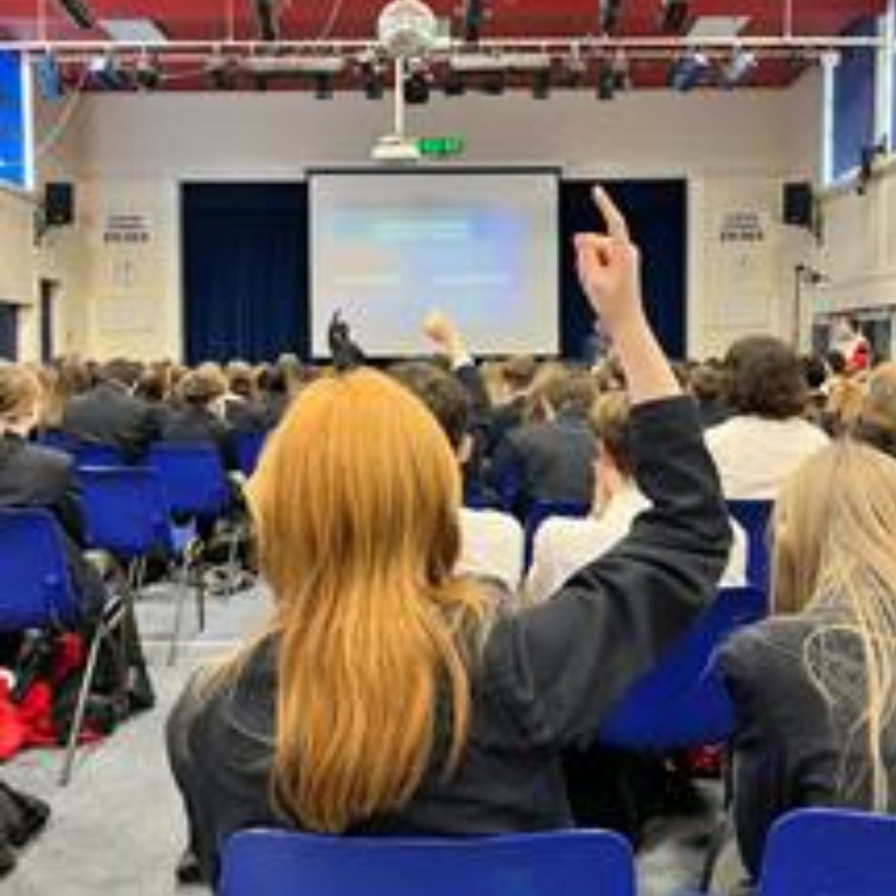 Young people seated in a school assembly hall during an educational presentation, with one student raising their hand to participate.