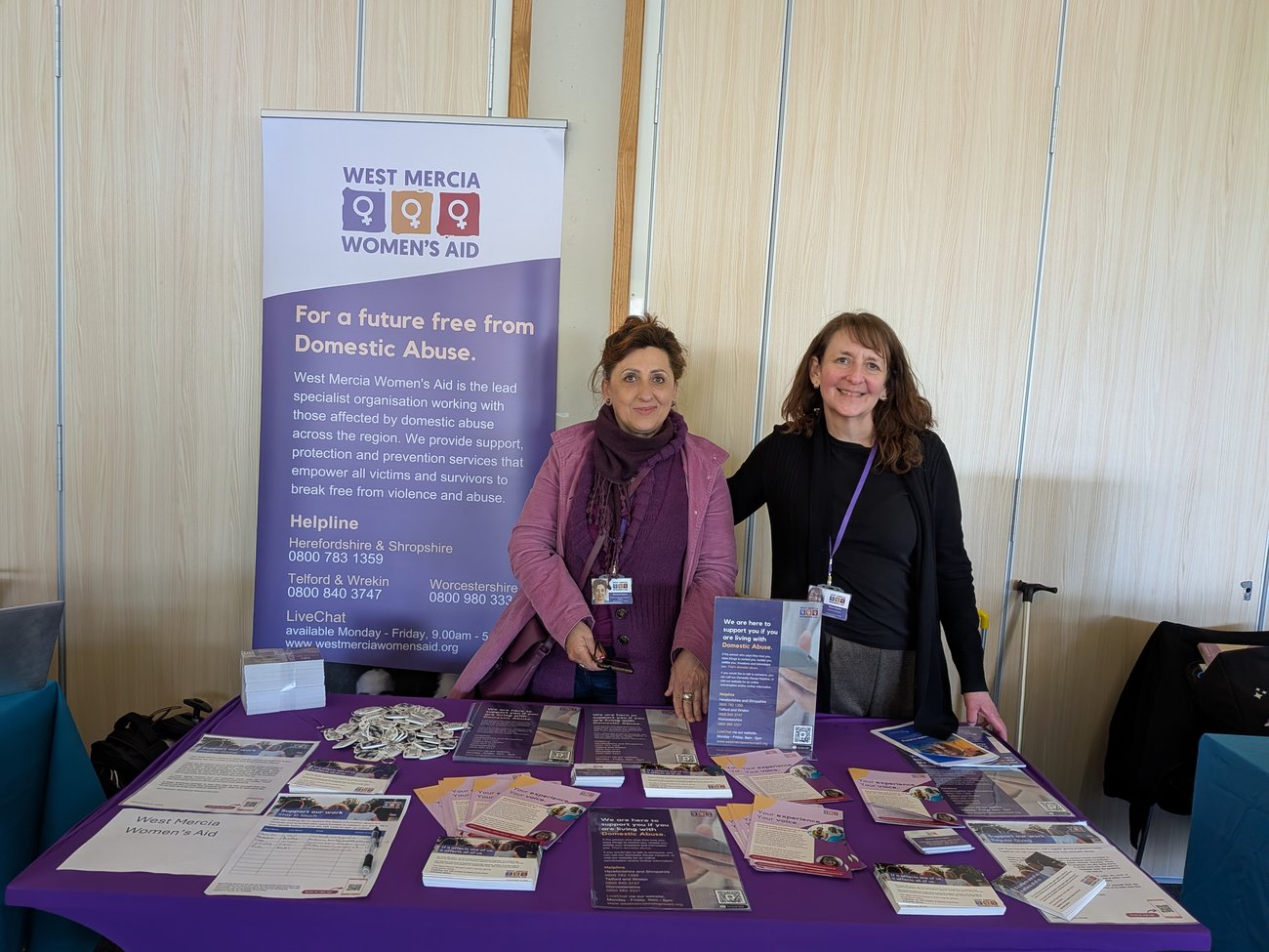Two West Mercia Women’s Aid staff members standing behind an information stall at a community wellbeing event, with leaflets and support materials on display.