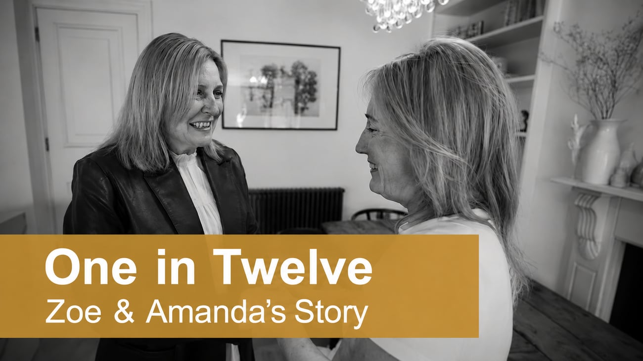 Black and white image of Zoe and Amanda smiling and holding hands in a living room, with a gold banner across the bottom reading “One in Twelve – Zoe & Amanda”.