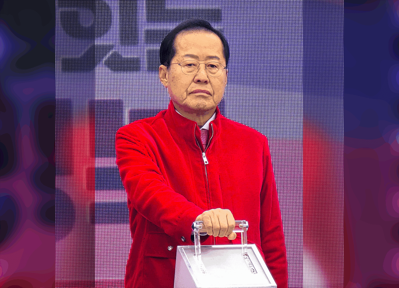 A man wearing glasses and a red jacket stands in front of a blurred background resting his hand on a metallic box