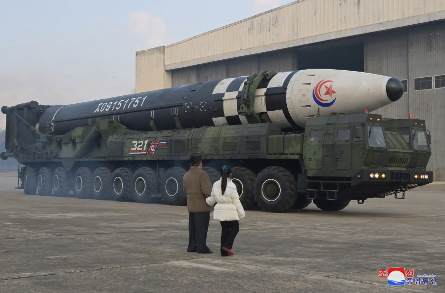 Kim Jong-un and his daughter facing away from camera look at very large missile on topof a very large long green military vehicle with many tires