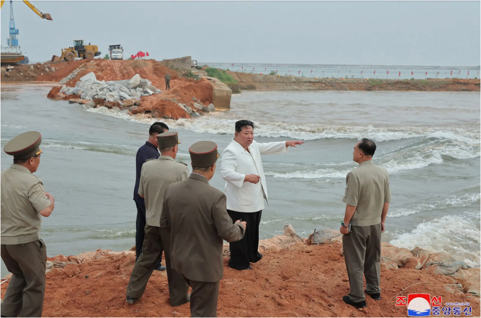 Kim Jong-un surveys an embankment that failed standing with other men in formal clothing looking at a fast-moving body of water damage to earthen wall in background