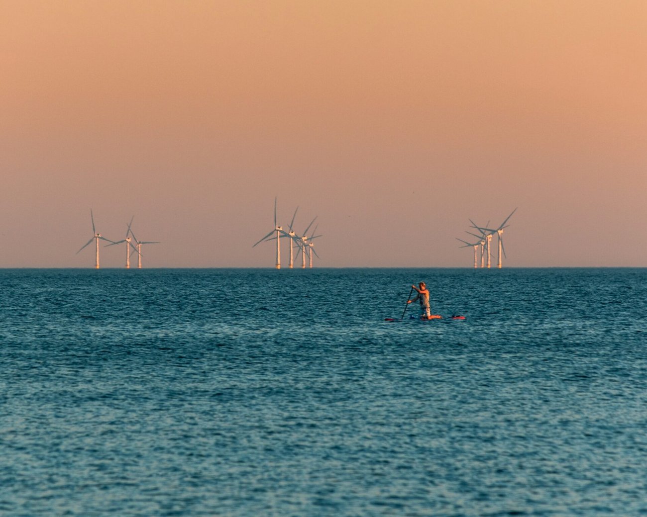 A paddleboarder paddles in ocean with wind turbines far in the background at either sunrise or sunset