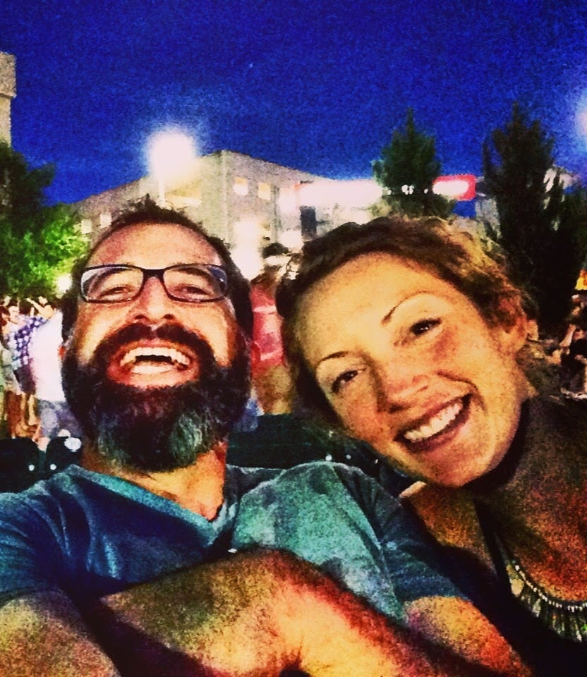 Abby and Andrew smiling together at an outdoor nighttime event, with lights and a crowd in the background.
