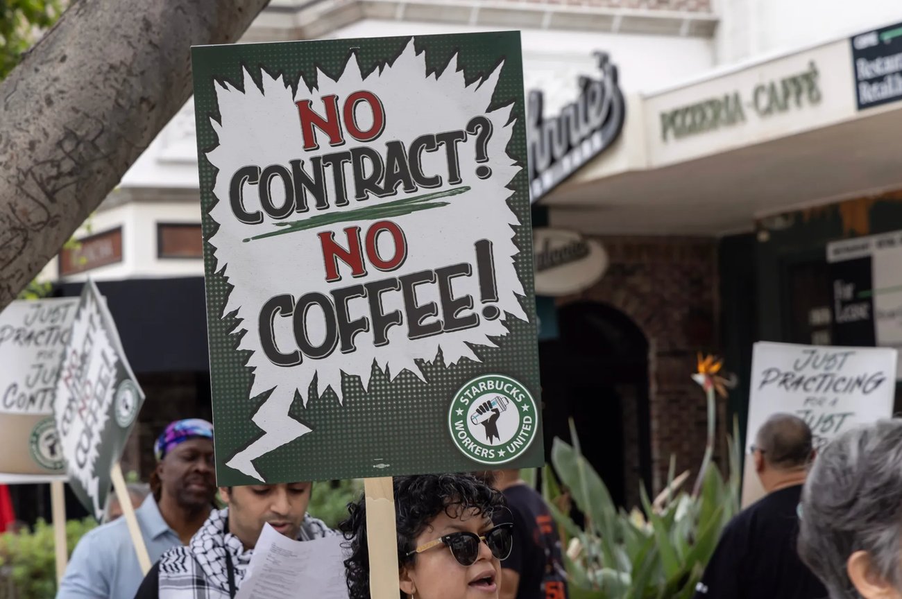 Protesters hold signs outdoors; the one in the center has a jagged speech bubble saying "NO CONTRACT? (underlined) NO COFFEE!" and has a small circular logo in the lower right corner, a fist holding a Starbucks cup encircled by "Starbucks Workers United".