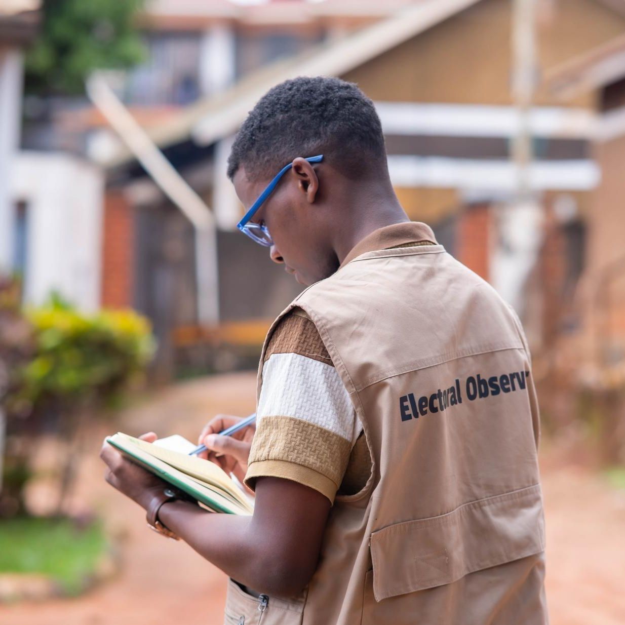 Isaac, wearing glasses and a tan vest marked "Electoral Observer", writes in a notebook on a residential street.