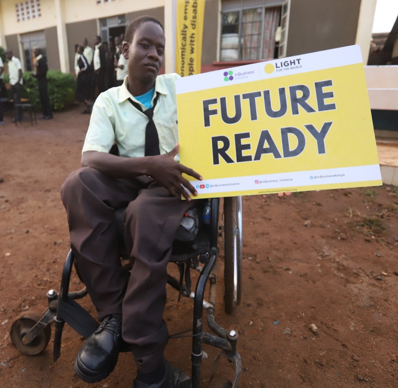 A student who uses a
wheelchair holds a yellow "Future Ready" sign carrying the InBusiness Initiative and Light for the World logos. He wears a school uniform; classmates stand in the courtyard behind him.