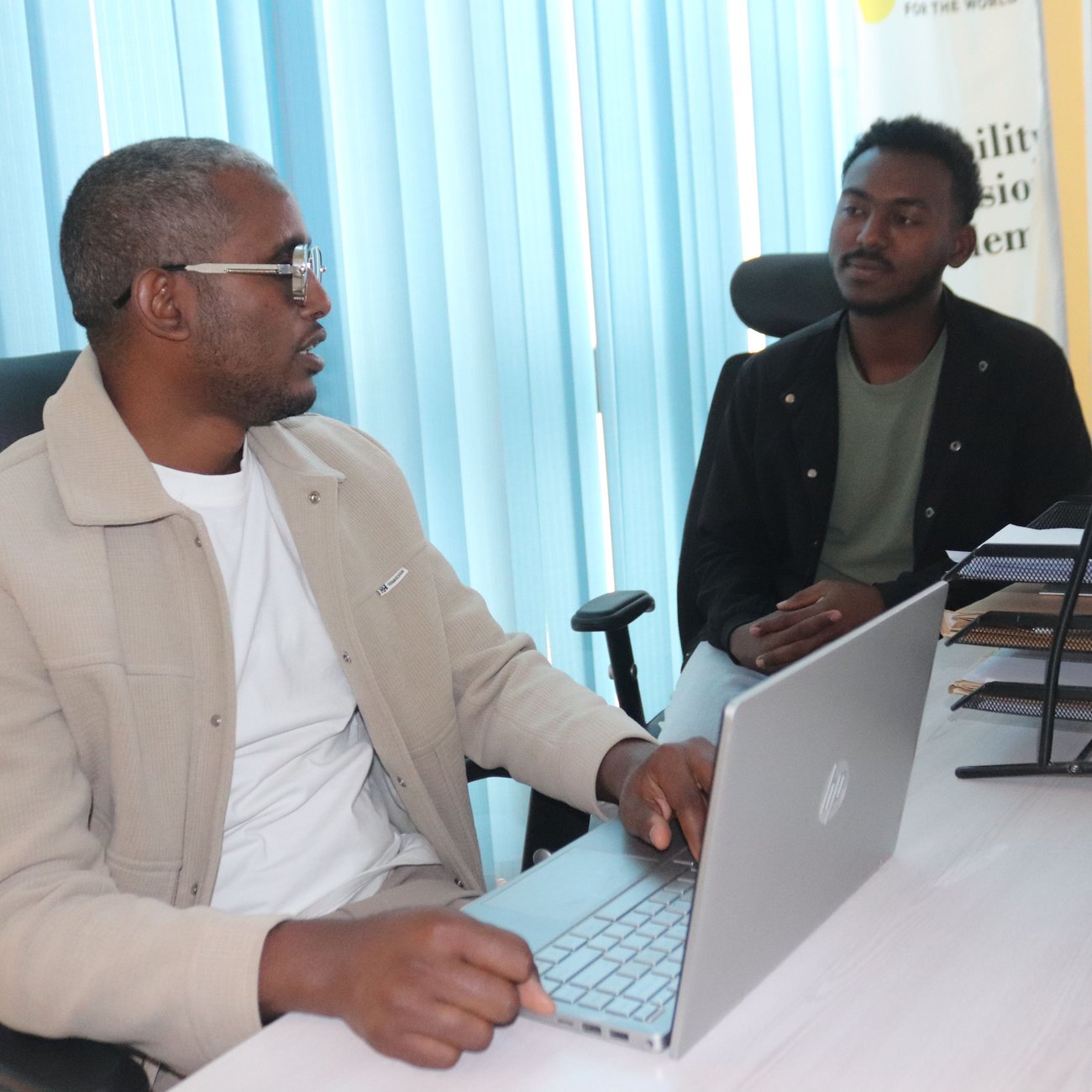 Yohannes sits at a desk in an office space, wearing sunglasses and a beige jacket, a laptop in front of him. He chats with a man to his left who wears a black shirt and a mint green t-shirt. 