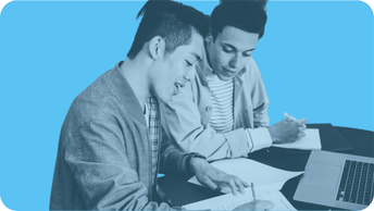 Two people sit next to eachother collaborating and writing on paper with a laptop nearby on the table. 