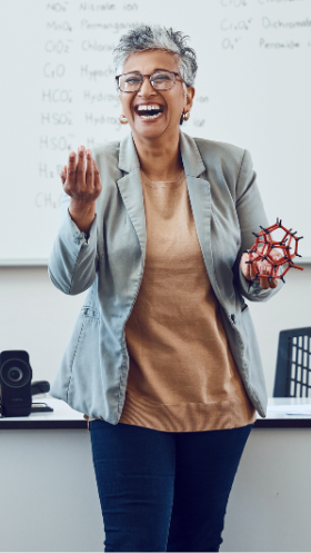 A
professor laughs while showing students a plastic model of a molecule in front of a classroom whiteboard. 