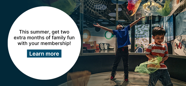 Two young children play in the ZOOOM - Children's Innovation Zone, surrounded by colorful, translucent fabric streamers. Overlaid on the left is a white circle with text that reads: "This summer, get two extra months of family fun with your membership! Learn more".