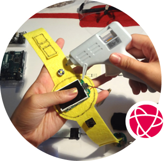 Two hands work with a glue gun and a yellow felt watch with electronic parts.