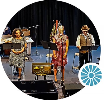 Three actors stand on stage behind sheet music holders and microphones.