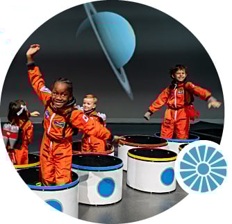 Smiling children wear orange space suit costumes and stand in colourful cylinders. A ringed image of a planet is visible in the background.