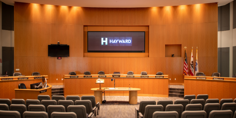 Photo of the City of Hayward's City Council Chamber. An auditorium with wood panelling, a wooden dais, audience chairs, and a large screen at the front. Photo of the City of Hayward's City Council Chamber. An auditorium with wood panelling, a wooden dais, audience chairs, and a large screen at the front.