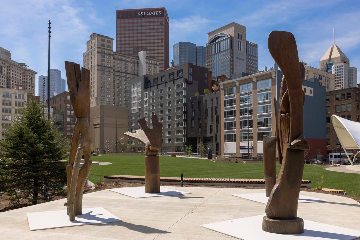 Sculptures of large abstract wooden hands are displayed in an urban park, with green grass and city skyscrapers in the background under a blue sky.