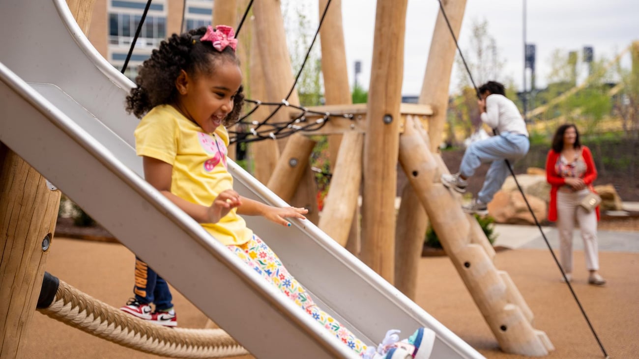 A young girl with curly hair and a pink bow smiles as she slides down a playground slide. In the background, another child climbs a rope structure while an adult stands nearby.