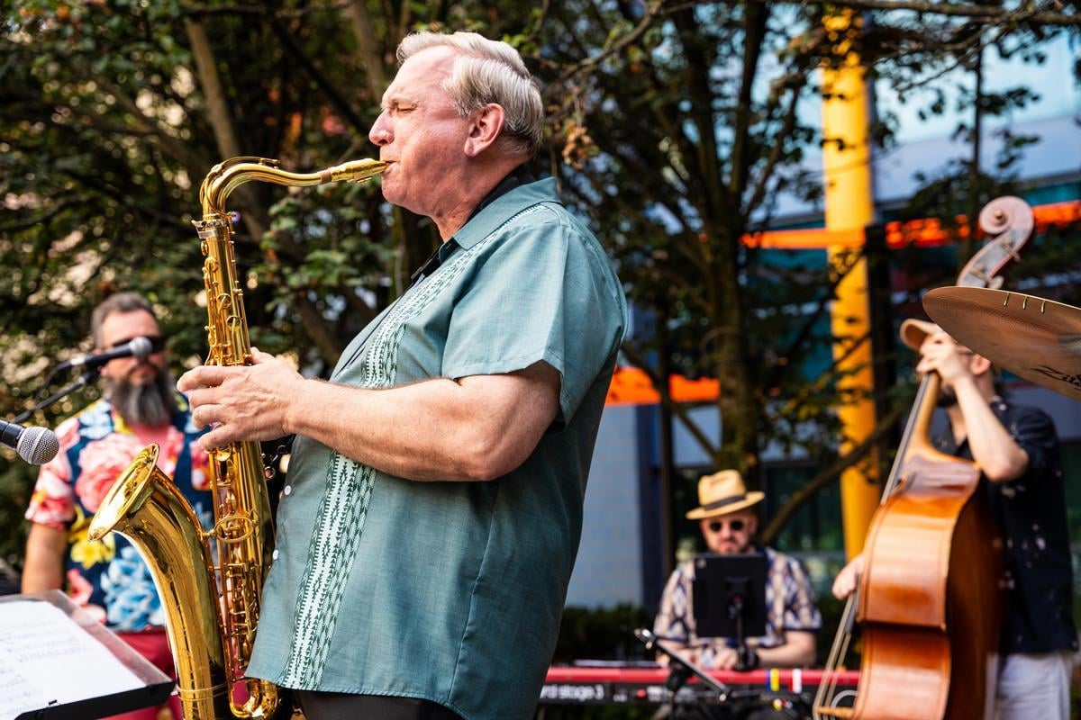A man in a green shirt plays the saxophone outdoors with a jazz band. A bassist, drummer, and another musician are visible in the background surrounded by trees and colorful structures.