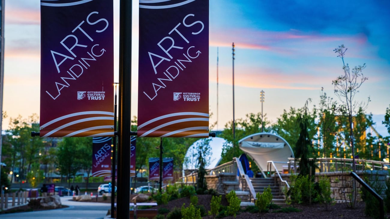 Banners reading Arts Landing hang on poles along a landscaped outdoor pathway at sunset, with a white tent structure and trees in the background.