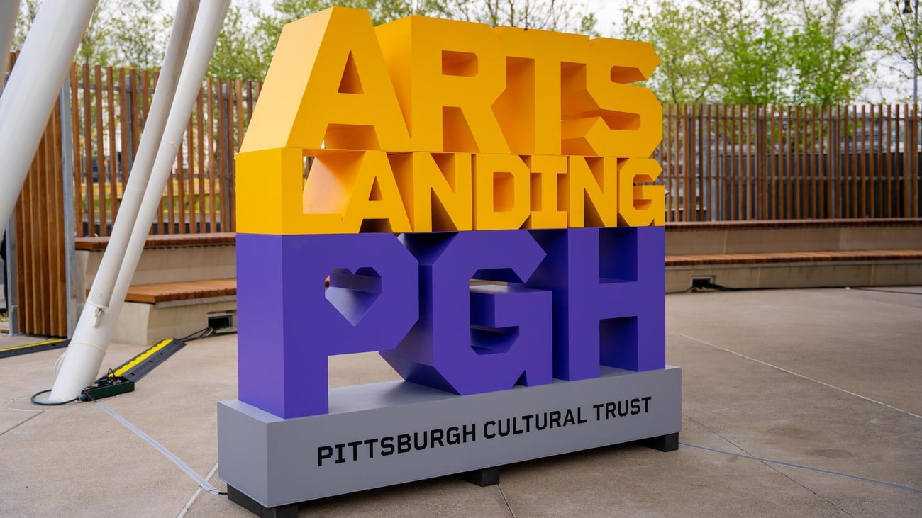 A colorful sign reads ARTS LANDING PGH in large yellow and purple letters, with Pittsburgh Cultural Trust written at the base. The sign is outdoors near a wooden fence and greenery.