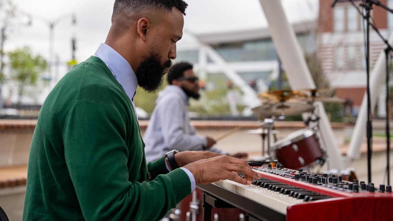 A man in a green sweater plays a keyboard outdoors while another musician in the background plays drums. Both are focused on their instruments, with modern buildings visible behind them.