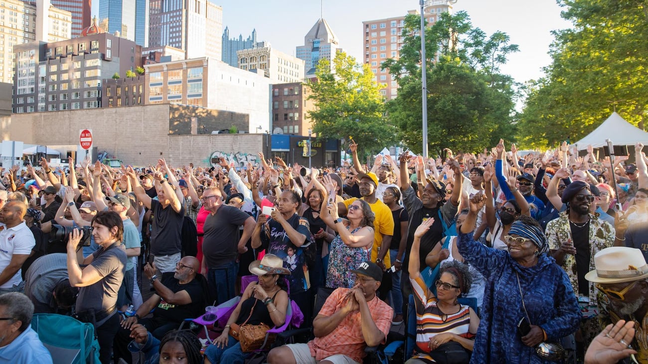 A large, diverse crowd of people stands outdoors in a city, many raising their hands enthusiastically. Tall buildings and trees are visible in the background under a clear sky.