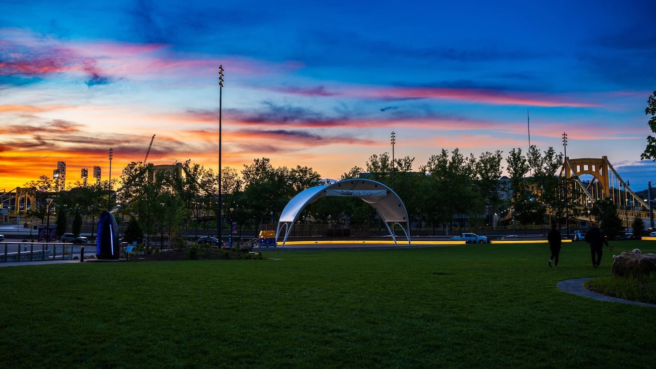 A park at sunset with a modern silver stage canopy, green lawn, and silhouetted trees. The sky is streaked with vibrant blue, orange, and pink clouds. Light poles and a distant bridge are visible in the background.