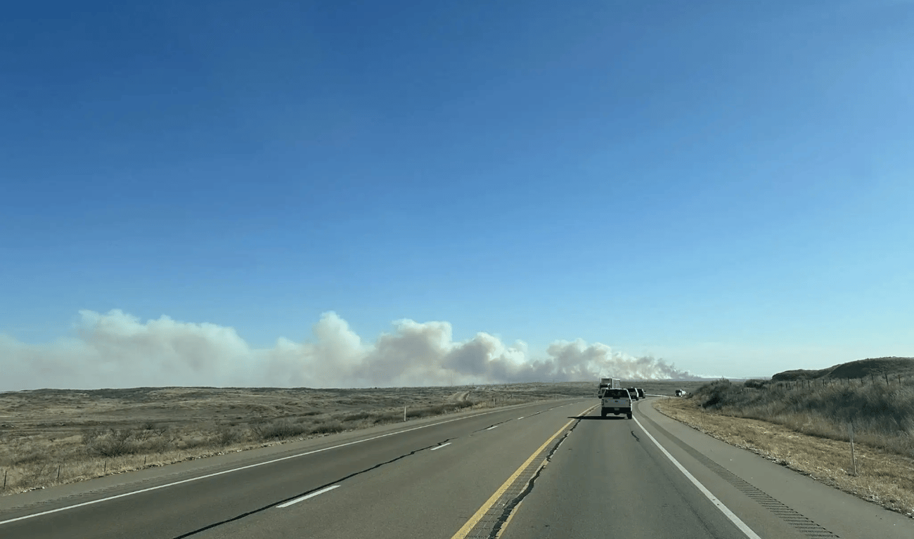 A wide-angle view from a vehicle traveling down a multi-lane highway toward a large, low-hanging plume of white and gray smoke on the horizon. The surrounding landscape consists of flat, dry brushland under a clear blue sky.