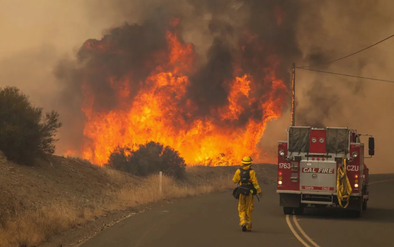 A firefighter in yellow protective gear walks along a paved road toward a massive wall of intense orange flames and thick black smoke. A red CAL FIRE engine is parked on the right side of the road, and the sky is a hazy, monochromatic orange.