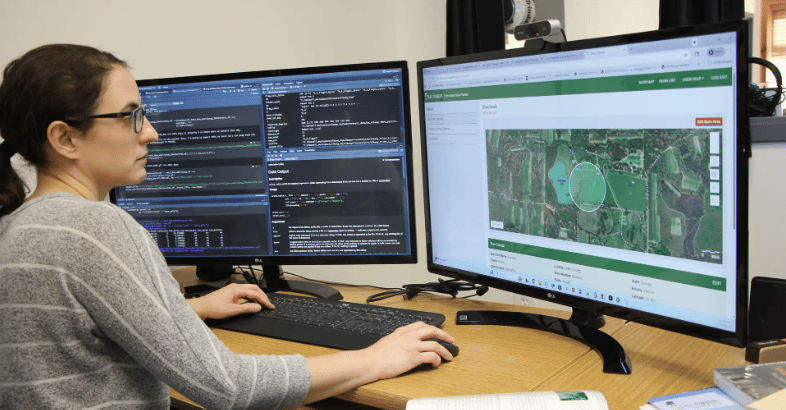 A woman with dark hair and glasses is seated at a wooden desk, working on two large computer monitors. She is wearing a grey striped sweater and using a black keyboard and mouse. She is demonstrated the Tall Timbers Prescribed Burn Planner. The monitor on the left displays code and technical documentation in a dark-themed interface, while the monitor on the right shows a mapping application with an aerial satellite view of a green landscape. A webcam is mounted on top of the right monitor. The desk also has a few books and papers scattered in the foreground.