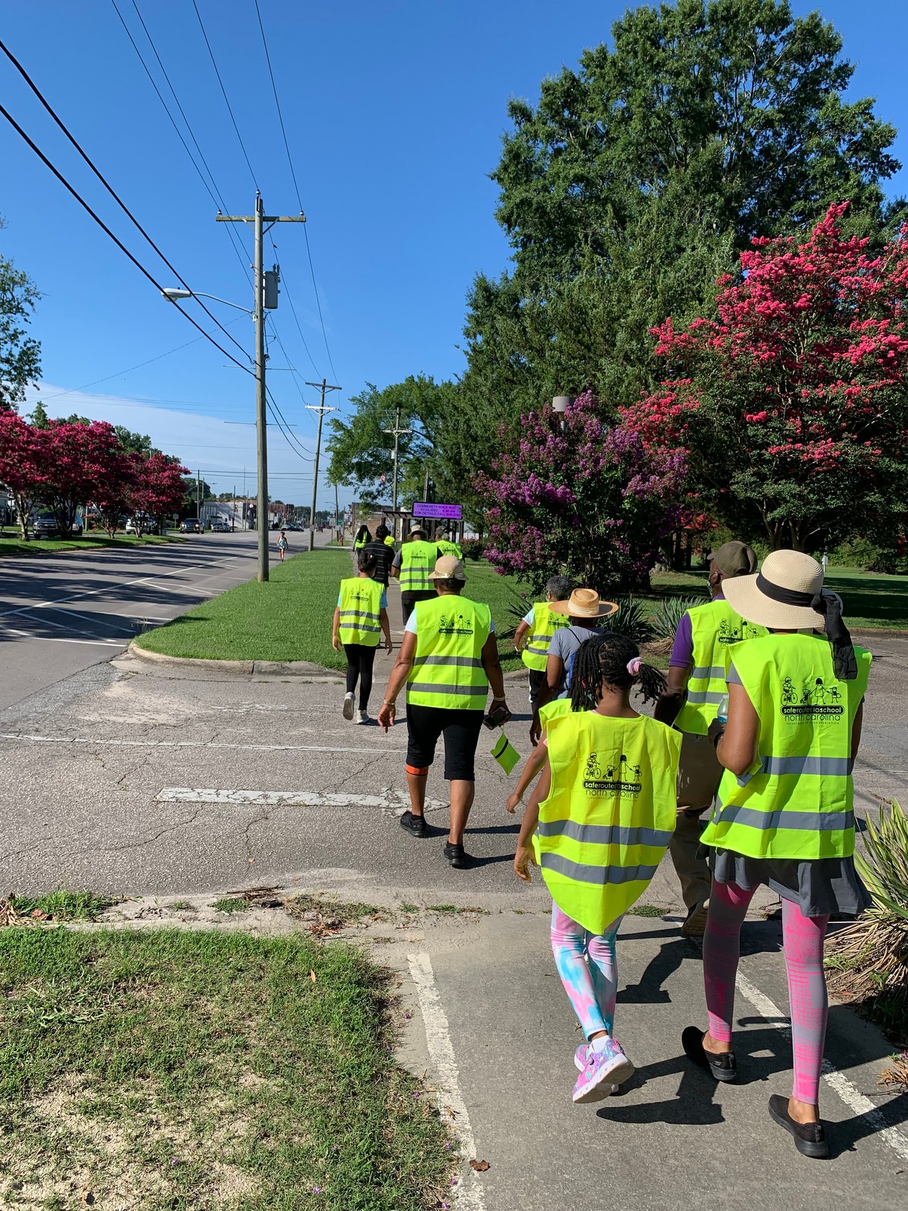 A group of adults and children wearing bright yellow safety vests walk along a sidewalk during a community walk audit, with a road, power lines, and blooming trees visible on a sunny day. A group of adults and children wearing bright yellow safety vests walk along a sidewalk during a community walk audit, with a road, power lines, and blooming trees visible on a sunny day.